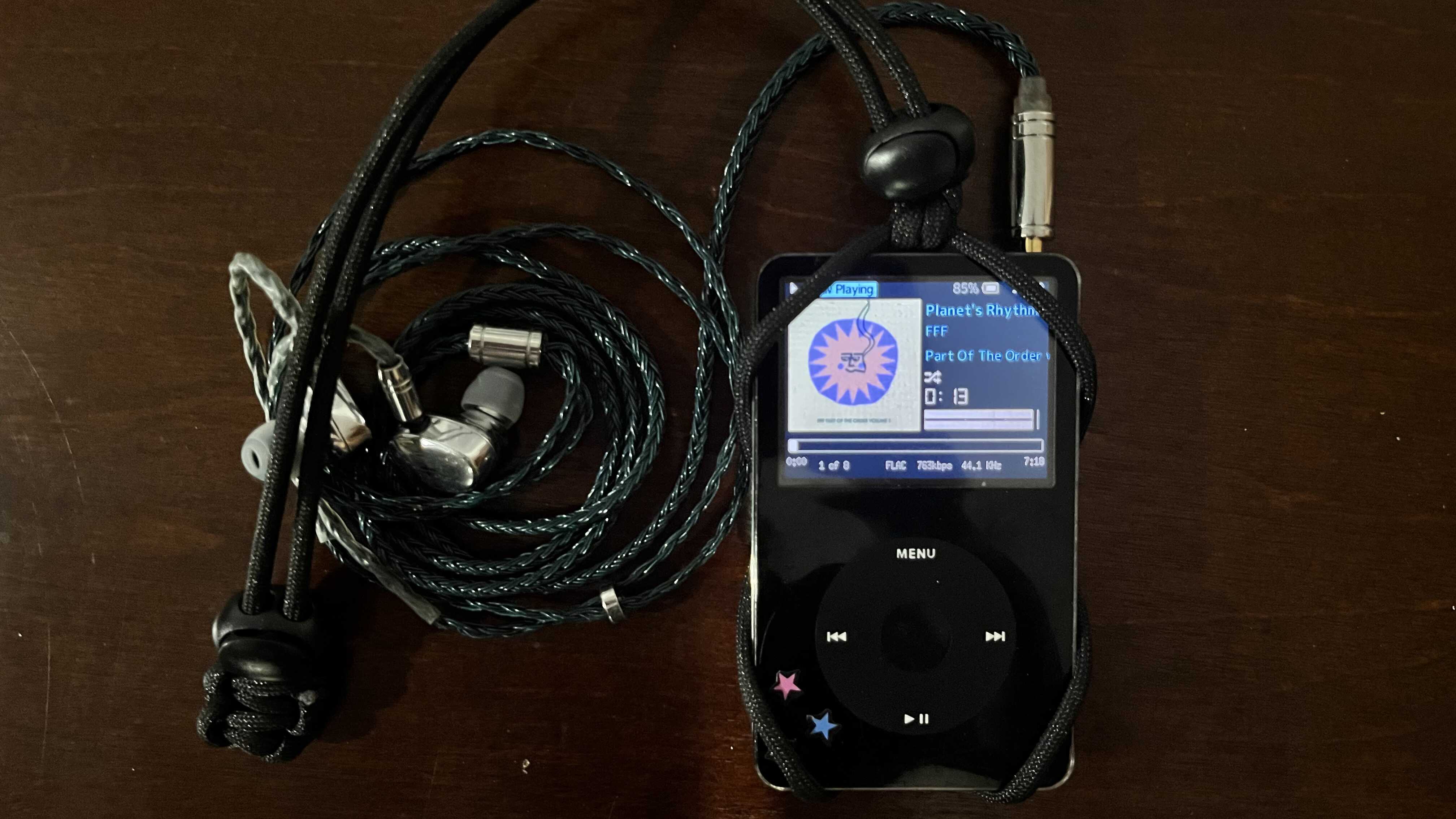 Black iPod Video with a paracord lanyard and earbuds. The iPod is on and it is displaying a now playing screen.